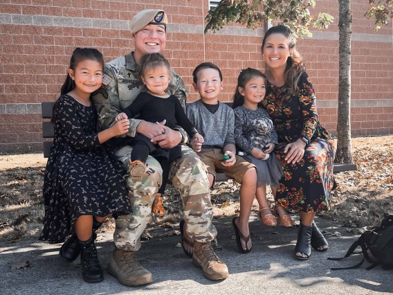 two parents and four children sitting on a bench. The father is wearing military camoflauge fatigues and a beret 